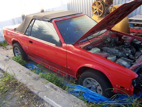 1984 Toyota Celica - A red convertible sports car sits in an outdoor lot with its hood open, showing the engine compartment.