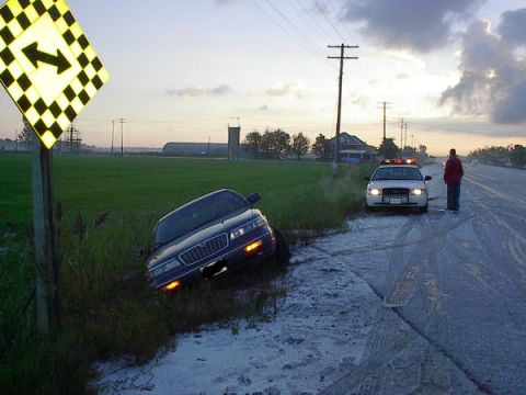1985 Mercury Grand Marquis - A dark sedan is in a ditch next to a highway, with a police car and an officer present at dusk.