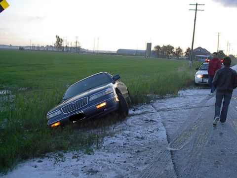 1985 Mercury Grand Marquis - A dark sedan is stuck in a ditch next to a rural road, with a police car and two onlookers present.