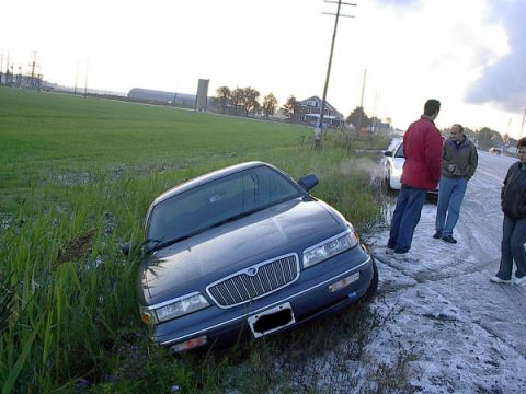 1985 Mercury Grand Marquis - A dark sedan is stuck in a ditch on the side of a wet road with three people observing.