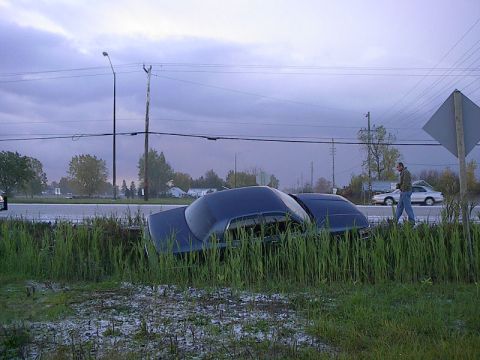 1985 Mercury Grand Marquis - A dark blue sedan is off the road and in tall grass, with a person walking nearby.