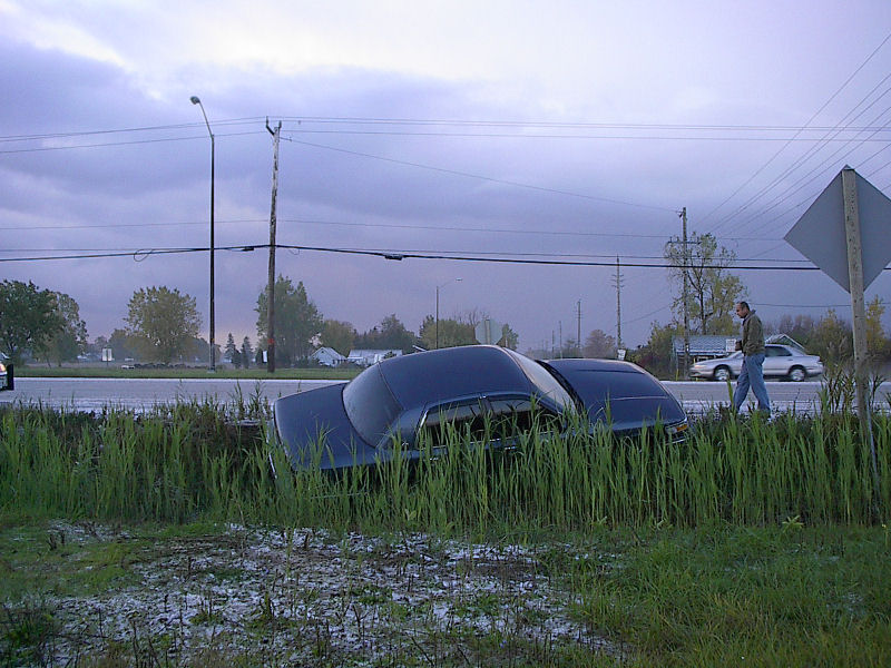 Dark blue sedan stuck nose-down in a flooded roadside ditch at dusk