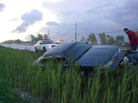 1985 Mercury Grand Marquis - A dark sedan is off the road in tall grass, with a police car responding to the scene.