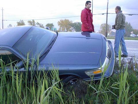 1985 Mercury Grand Marquis - A blue sedan is stuck in tall grass and mud next to a road, with two men standing nearby.