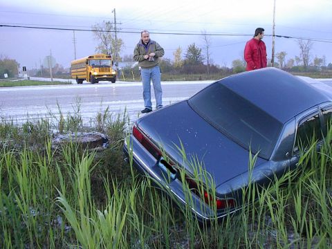 1985 Mercury Grand Marquis - A dark sedan is off the road in tall grass, with two men standing nearby and a school bus in the background.