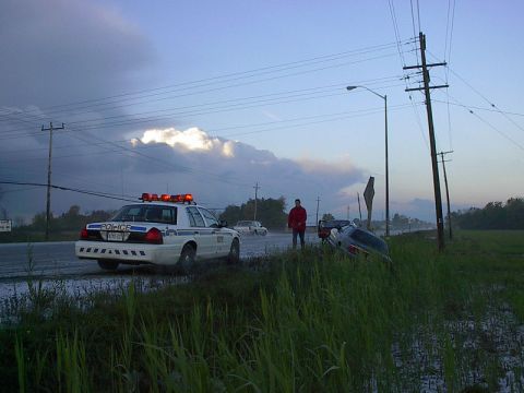 1985 Mercury Grand Marquis - A police car is present at a scene where another vehicle has crashed into a ditch, with a person standing nearby.