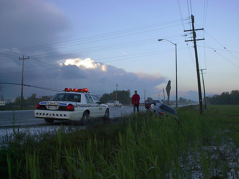 White OPP Crown Victoria cruiser on wet highway beside dark sedan stuck in flooded ditch