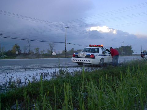 1985 Mercury Grand Marquis - A police car is parked on the side of a wet road under a cloudy sky, with a person leaning into the driver's side window.