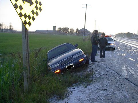 1985 Mercury Grand Marquis - A dark sedan is off the road with a police car and two individuals nearby.