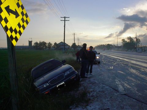 1985 Mercury Grand Marquis - A dark sedan is off the road near a curve sign, with a police car and onlookers present.
