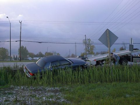 1985 Mercury Grand Marquis - A tow truck is preparing to load a dark sedan that has driven off the road into tall grass.