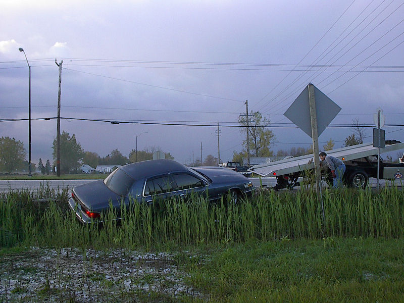 Dark blue sedan stuck in a grassy roadside ditch with a flatbed tow truck tilting its deck to recover it