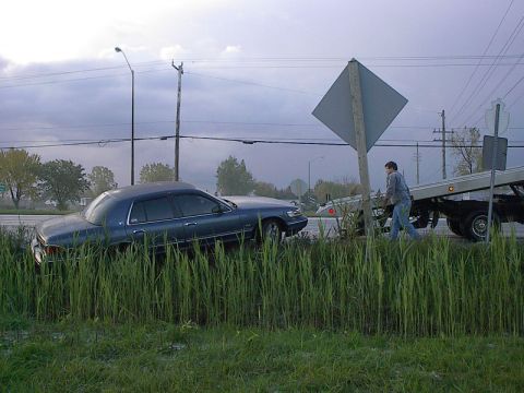 1985 Mercury Grand Marquis - A blue sedan is being loaded onto a tow truck after going off the road into tall grass.