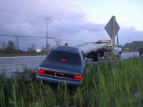 1985 Mercury Grand Marquis - A dark blue sedan is stuck in a ditch beside a road, being loaded onto a flatbed tow truck.