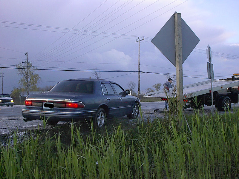 Blue-gray Mercury Grand Marquis on a roadside shoulder with a flatbed tow truck deck tilted behind