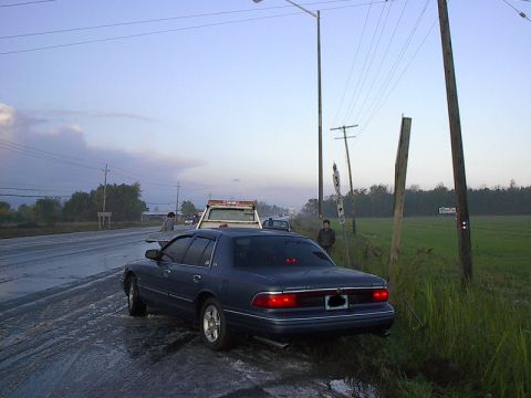1985 Mercury Grand Marquis - A dark blue sedan, likely a 1996 Mercury Grand Marquis, is pulled over on a wet road with a tow truck visible behind it.