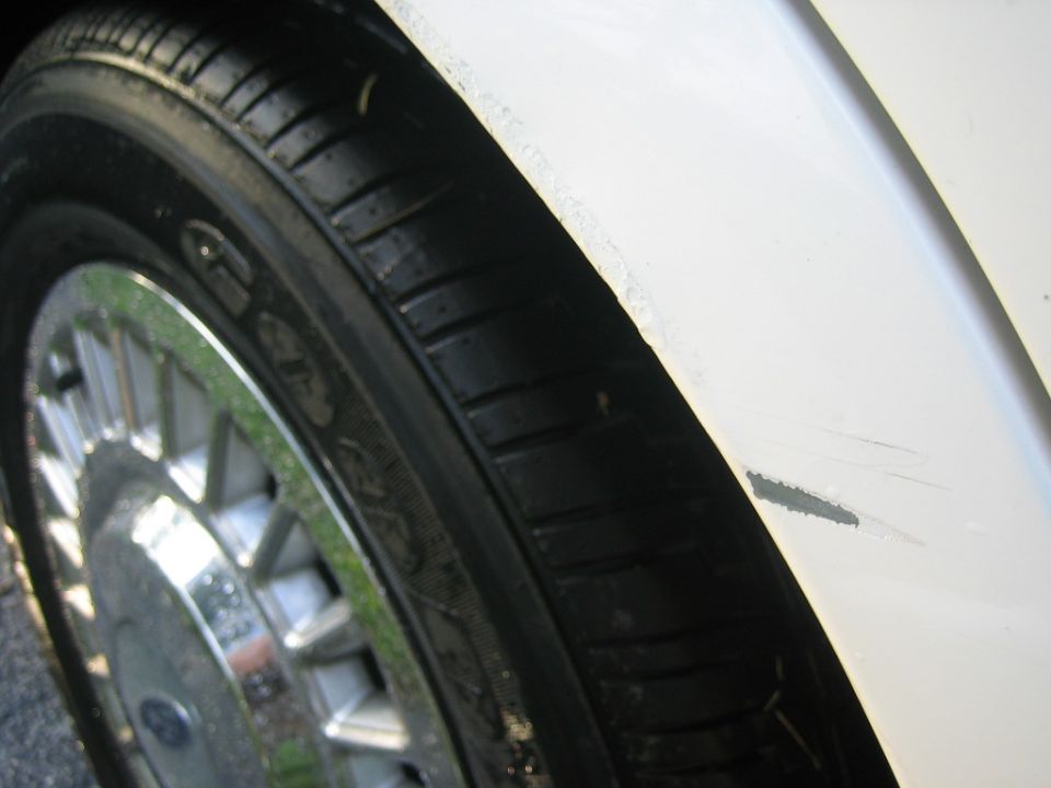 Close-up of paint chips and scratches on white fender above tire with chrome wheel cover