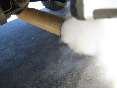 1990 Ford Bronco - Thick white smoke pours from a tan exhaust pipe protruding beneath a lifted vehicle parked on asphalt.