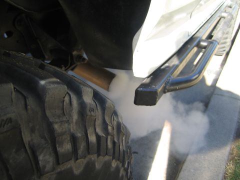 1990 Ford Bronco - White exhaust smoke escapes from beneath a vehicle next to a chunky off-road tire and a black tubular side step.
