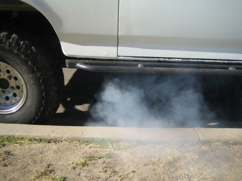 1990 Ford Bronco - White exhaust smoke billows from under the rocker panel of a white truck with an off-road tire and tubular side step over a curb.