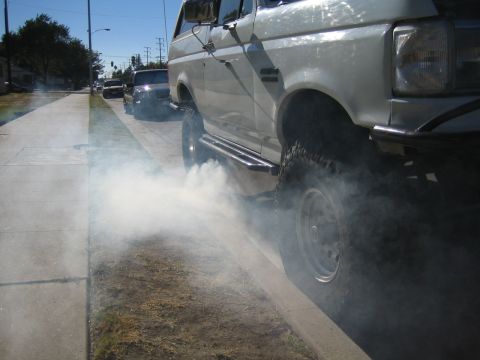 1990 Ford Bronco - white lifted SUV with off-road tires emits heavy white smoke at its front wheel while parked along a curb on a sunny street.