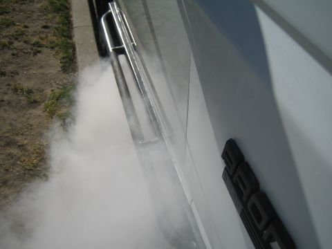 1990 Ford Bronco - Angled view of a white vehicle's body and chrome tube side step with white smoke rolling out below over pavement.