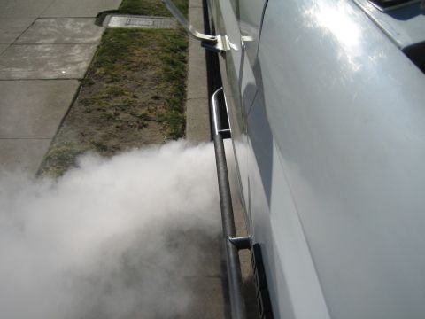 1990 Ford Bronco - White smoke pours from beneath the side of a white vehicle with a chrome tube step, drifting across a sidewalk and grass strip.