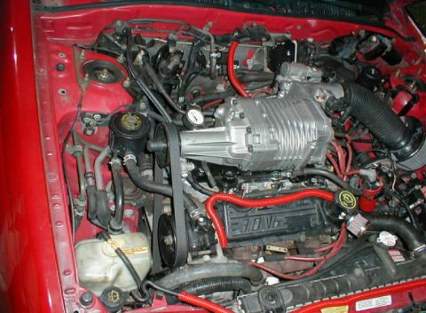 1992 Ford Probe - Engine bay of a red car fitted with a supercharger conversion on a transverse V6, with a Roots-type blower and intake plenum mounted above…