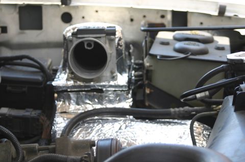 1994 Ford Bronco - Close-up of an engine bay component wrapped in reflective silver heat-shield insulation, with a large round open duct or air intake opening…