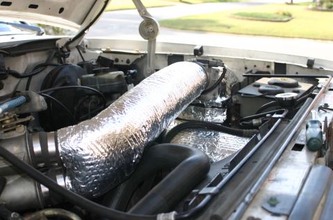 1994 Ford Bronco - Engine bay view of a vehicle with a large intake or air tube wrapped in silver reflective heat-insulation, alongside black hoses, a battery…