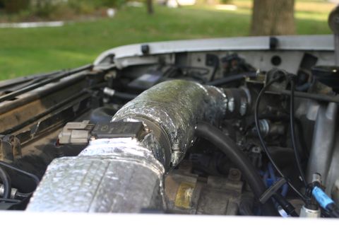 1994 Ford Bronco - Close-up of an engine bay showing intake tubing and a component wrapped in reflective foil heat-shielding insulation, with black vacuum hos…