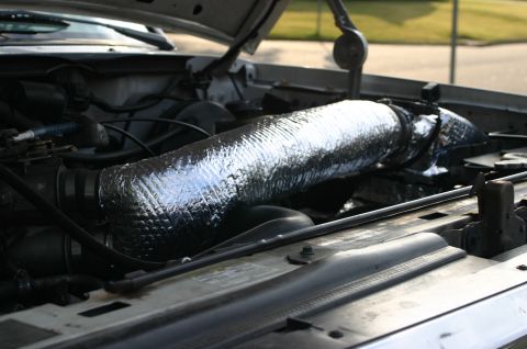 1994 Ford Bronco - Engine bay of a 1994 Ford Bronco with the hood raised, showing a large intake tube wrapped in reflective silver heat-insulating material. N…