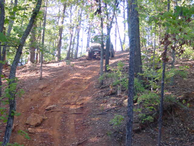 Distant Jeep cresting a steep red-clay trail surrounded by tall trees in a forest