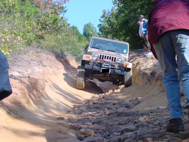 Jeep TJ going down a rocky, rutted dirt trail with trees and people around