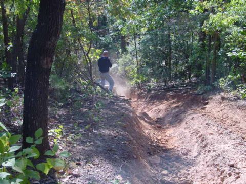 2004 Jeep Wrangler - A person stands beside a dusty off-road trail carved through a wooded area.