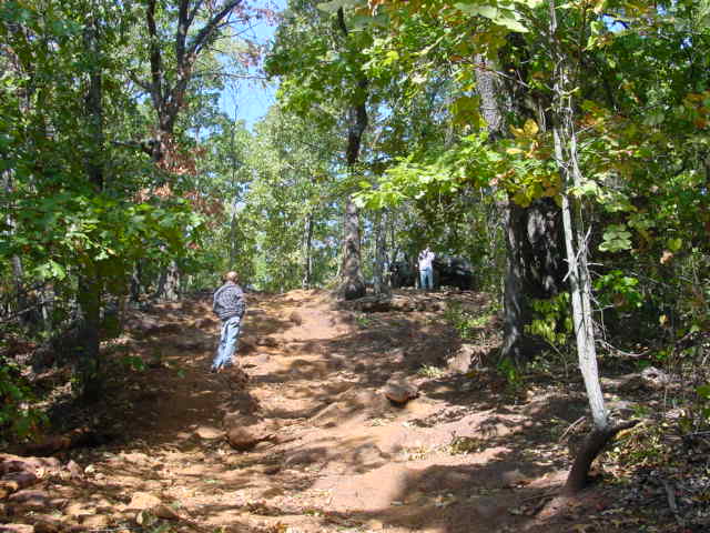 Wooded trail scene with two people and a Jeep in the distance