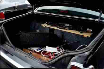 1991 Ford LTD Crown Victoria - Open trunk of a dark sedan showing carpeted interior, a board with mounted electronics, and red and white wiring.
