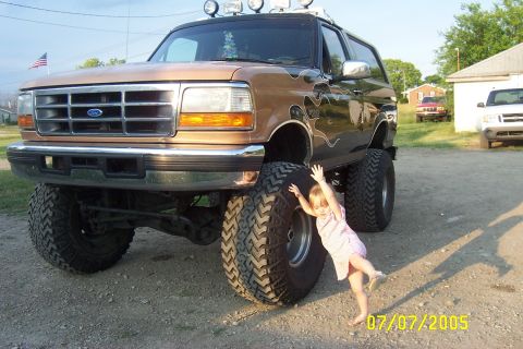 Brown lifted Ford Bronco with flame decals and large tires, a child is posing next to the front tire