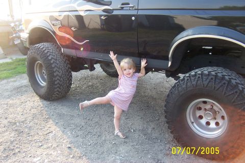 Kid posing under lifted black Ford Bronco with flame decals. Bronco badge visible on fender