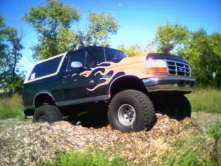 Black and tan Ford Bronco with flame decals, lifted on large tires, parked on a pile of wood chips