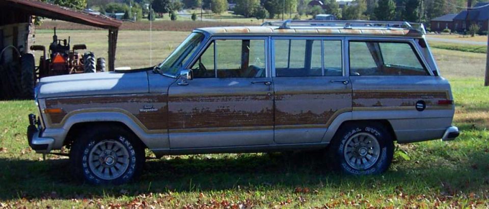 Silver Jeep Grand Wagoneer side view with peeling woodgrain trim, parked on grass