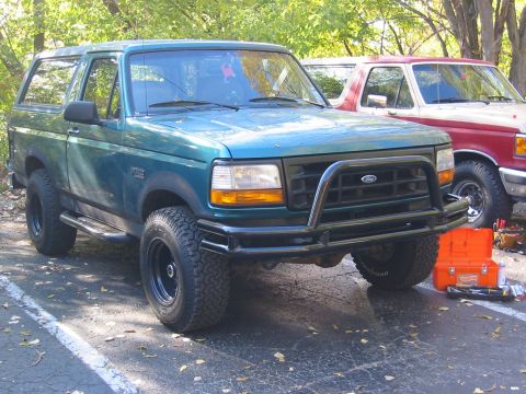 1996 Ford Bronco - A green 1996 Ford Bronco is parked next to a red truck, equipped with a black bull bar on the front bumper.