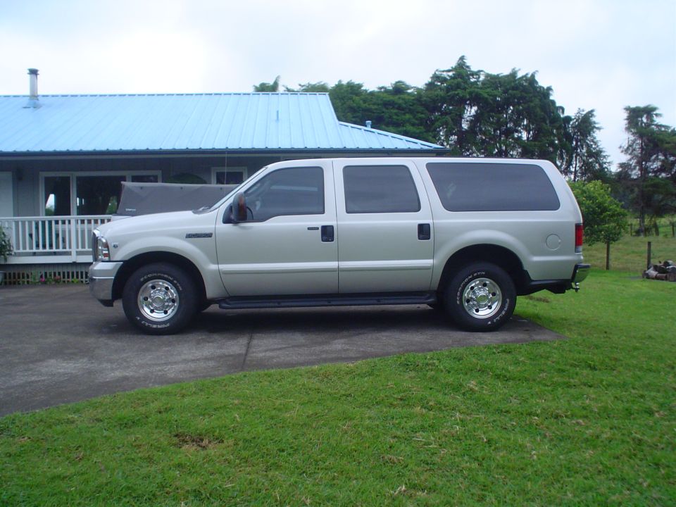 Side view of a silver Ford Excursion SUV parked on a driveway by a house with a blue roof