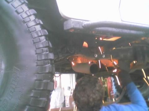 1990 Ford Bronco - person welds under a raised vehicle near the exhaust pipe, sending sparks flying beside a large mud-terrain tire.