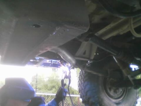 1990 Ford Bronco - Underbody view of a lifted vehicle showing the skid plate, exhaust routing and off-road tire, with a welding glow at the lower edge.