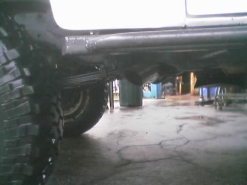 1990 Ford Bronco - Side underbody view of a lifted truck showing a leaf spring, exhaust pipe and off-road tire over a cracked concrete shop floor.
