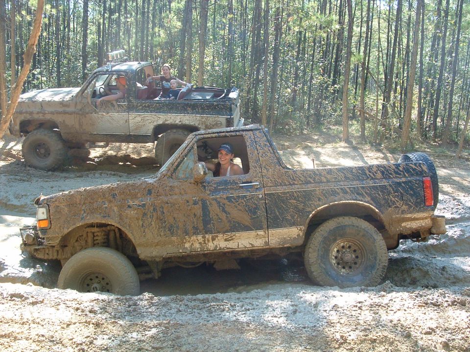 Muddy topless Ford pickup stuck in deep mud with a person inside, second truck behind in pine woods