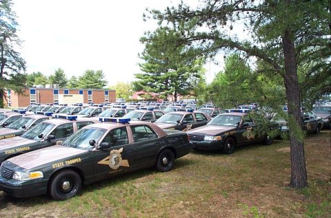 1991 Ford LTD Crown Victoria - A large fleet of police vehicles, likely from the same department, are parked outdoors under trees.