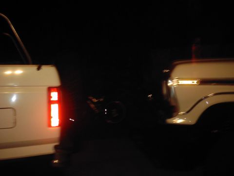 1978 Ford Bronco - Two white trucks are visible at night, with the rear of a 1978 Ford Bronco illuminated by its brake light.