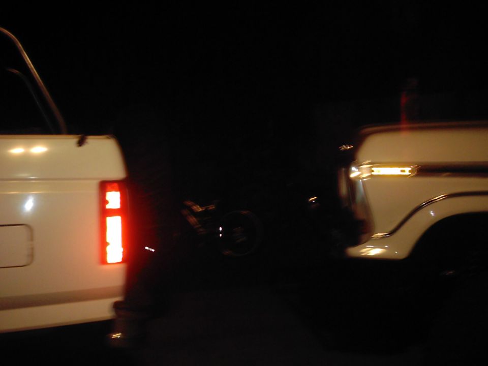 Rear of a white 1978 Ford Bronco with its brake light on, next to another white truck
